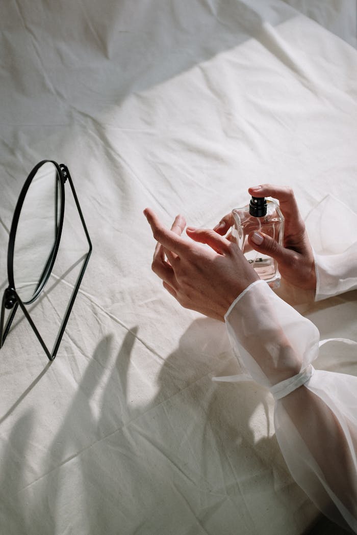 Delicate hands holding a perfume bottle beside a mirror on a white cloth background.